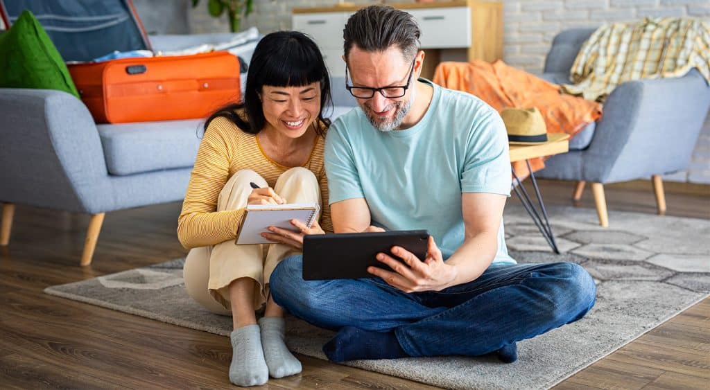 A couple is getting ready for a trip; a woman is writing a checklist, and a man is using a digital tablet
