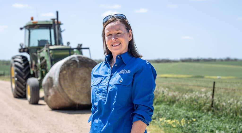 Katrina Nankivell standing on her Maitland farm smiling at the camera with a tractor and wheat fields in the background.
