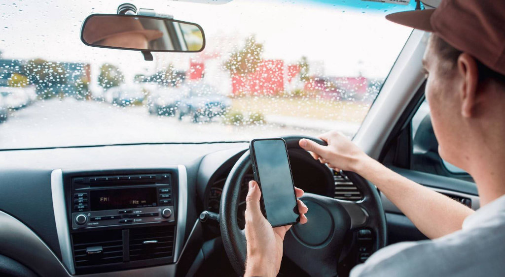 A young man driving with one hand while also using his phone in the other hand.