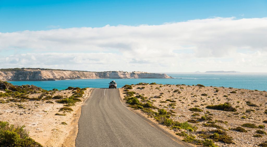 A vehicle drives along a sealed road in Coffin Bay National Park with an ocean landscape in the background