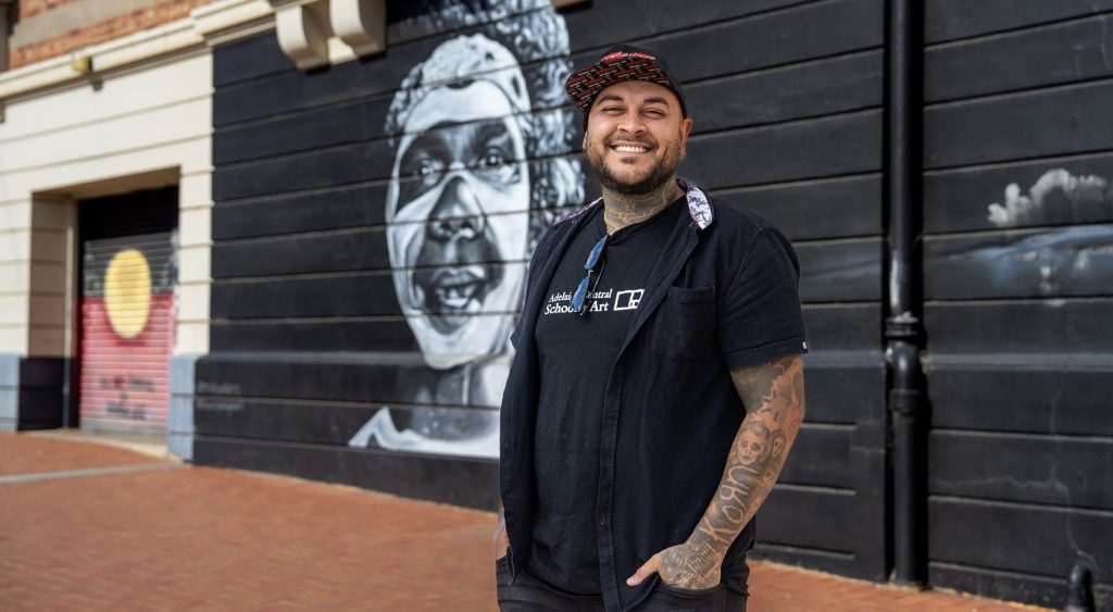 Thomas Readett standing in front of mural of well-known Indigenous actor David Gulpilil.