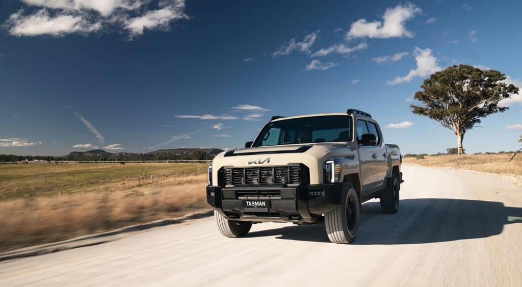 Kia Tasman ute driving on a rural gravel road under a blue sky with scattered clouds.