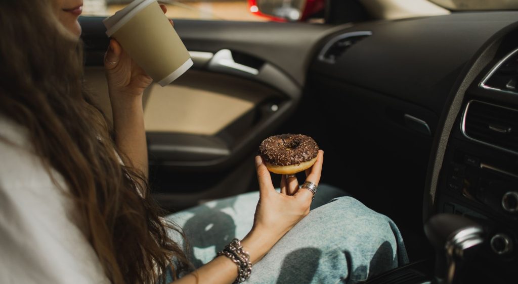 Woman eating doughnut and drinking coffee in the passenger seat of a car.