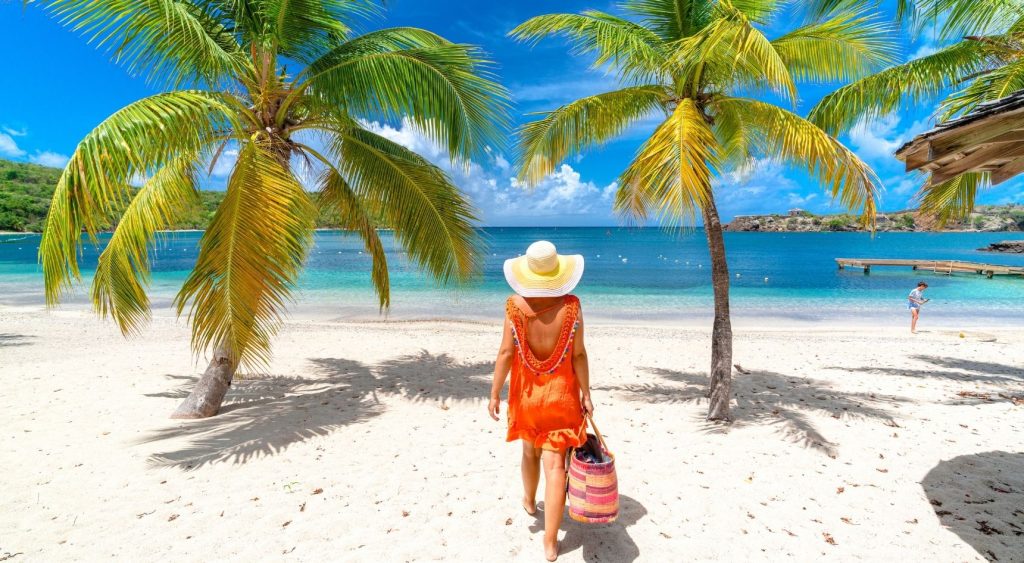 Woman walks on a tropical island, wearing a wide-brimmed hat and carrying a beach bag.